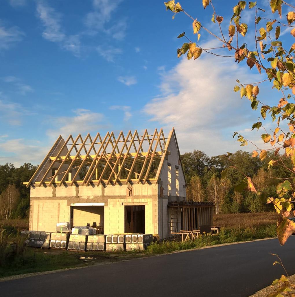 Surowy stan budowy domu jednorodzinnego z bloczków, widoczne drewniane krokwie dachu, palety z materiałem budowlanym oraz jesienne liście na drzewie w tle.