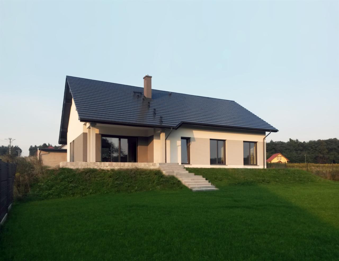 Nowoczesny dom jednorodzinny z ciemną dachówką i dużymi oknami, otoczony zielonym trawnikiem, widok z przodu, lekko pochmurne niebo.