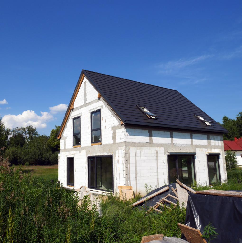 Surowy stan domu jednorodzinnego z czarnym dachem i oknami, widoczne białe bloczki, nieotynkowana elewacja i materiały budowlane wokół.