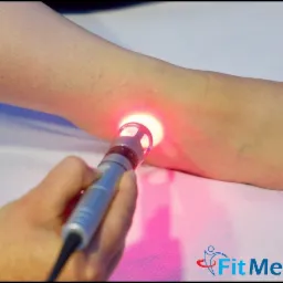 Laseroterapia na stopie pacjenta z użyciem urządzenia z metalową obudową i czerwonym światłem, na tle białego prześcieradła, widoczne logo FitMed.