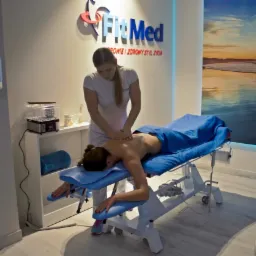 Kobieta w białym uniformie wykonuje masaż pleców pacjentki leżącej na niebieskim łóżku do masażu w jasnym pomieszczeniu z logo firmy FitMed na ścianie i obrazem plaży w tle.