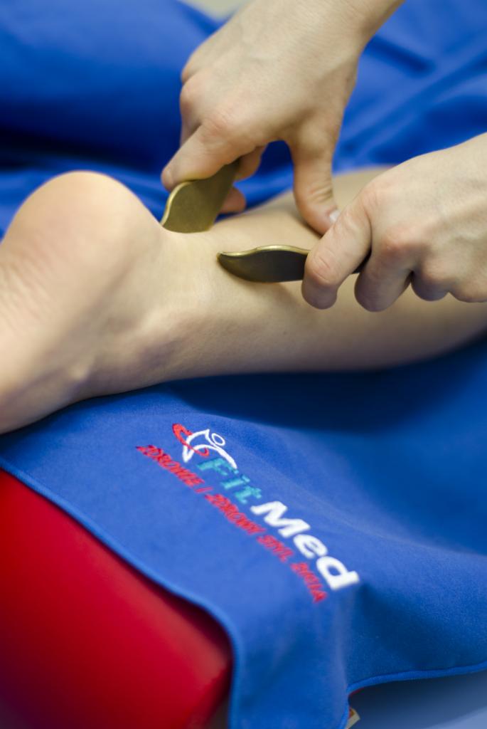 Dłonie terapeuty używają dwóch gładkich, metalowych narzędzi do masażu tkanek miękkich na łydce pacjenta, na niebieskim podkładzie z logo 'Fit Med'.