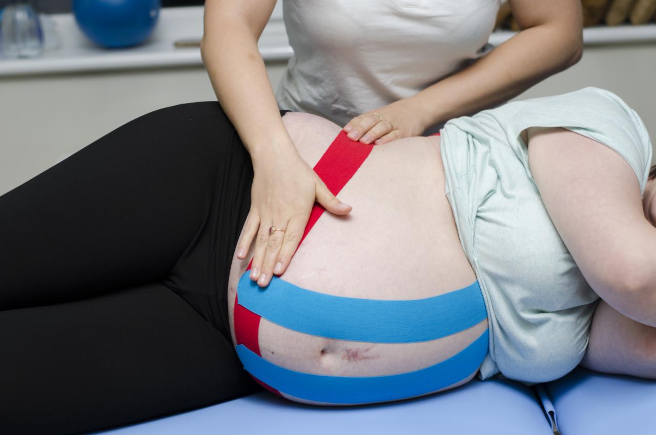 Kobieta w ciąży leżąca na boku z aplikacją taśm kinesiotaping w kolorach czerwonym i niebieskim na brzuchu, dłoń terapeuty delikatnie dotyka taśmy.