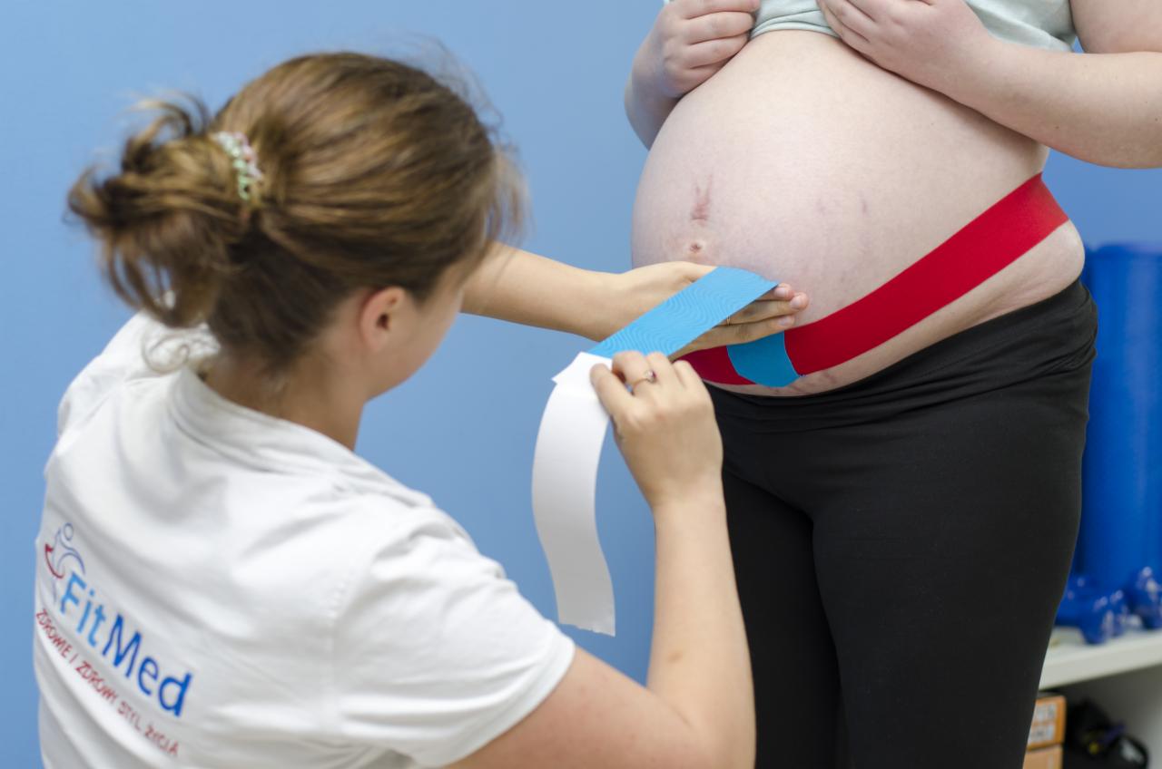 Aplikacja kolorowych taśm kinesiotaping na brzuchu kobiety w ciąży przez specjalistę w białym uniformie z logo FitMed na plecach.