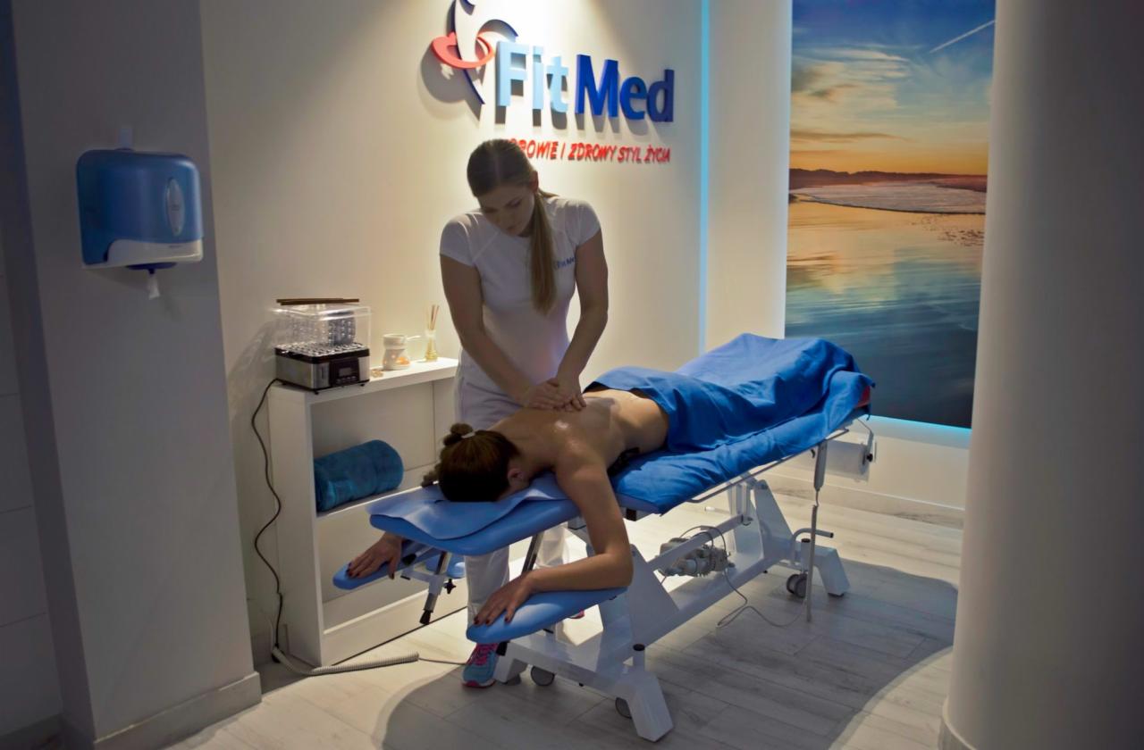 Kobieta w białym uniformie wykonuje masaż pleców pacjentki leżącej na niebieskim łóżku do masażu w jasnym pomieszczeniu z logo firmy FitMed na ścianie i obrazem plaży w tle.