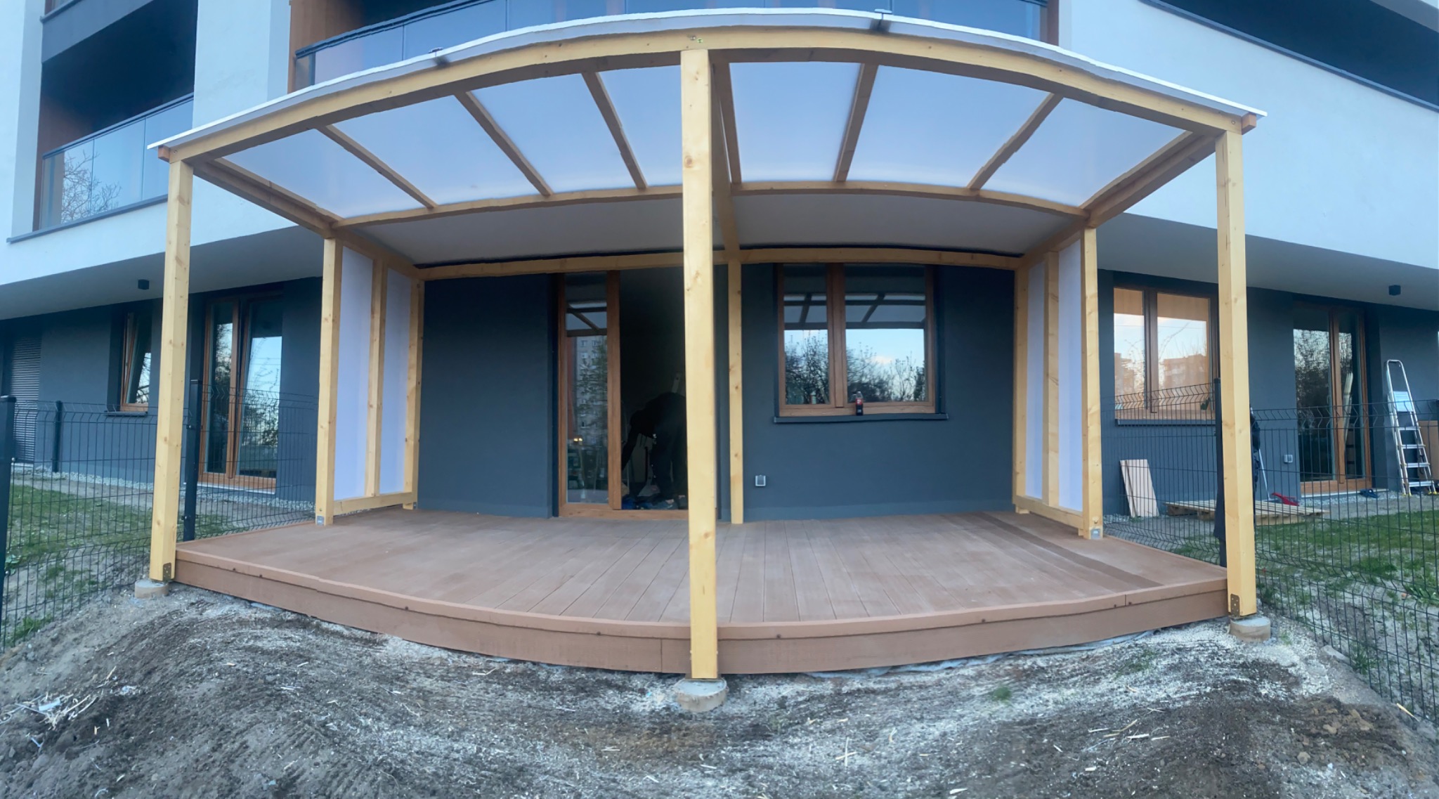 Zadaszony taras kompozytowy z drewnianą konstrukcją, półprzezroczystym dachem i widocznymi elementami montażowymi, umiejscowiony przy nowoczesnym budynku mieszkalnym.