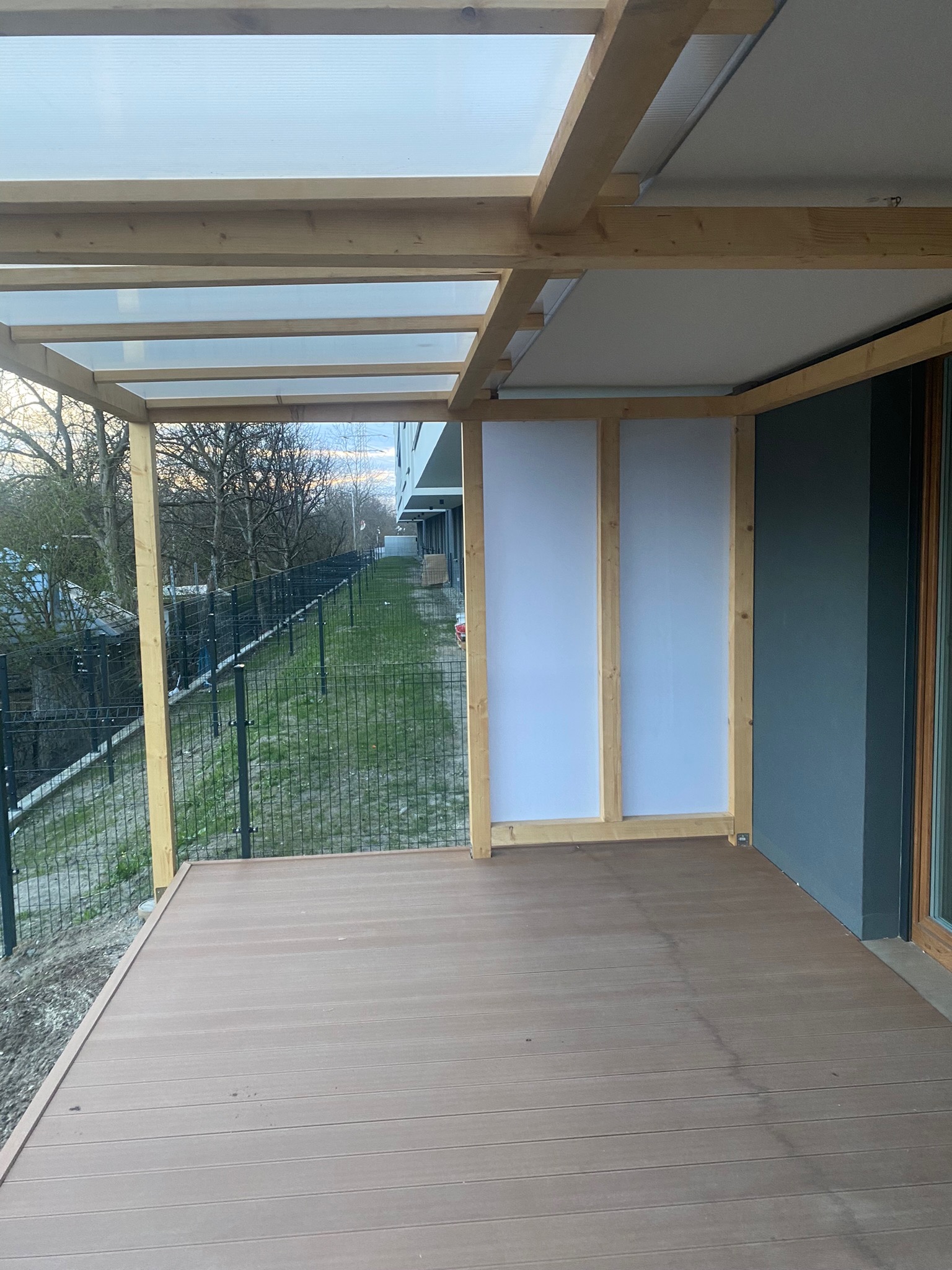 Zadaszony taras kompozytowy z drewnianą konstrukcją, częściowo osłonięty białymi panelami, widok na ogrodzony teren zielony.