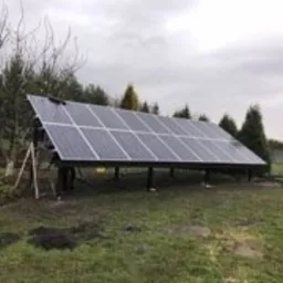 Nisko zawieszona instalacja paneli słonecznych na metalowej konstrukcji, widoczna osoba pracująca przy panelach, trawnik i drzewa w tle, pochmurne niebo.