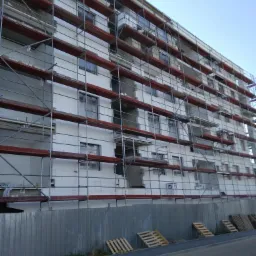 Wielopiętrowy budynek w trakcie ocieplania, pokryty rusztowaniami i częściowo płytami izolacyjnymi, widoczne drewniane palety opierające się o blaszane ogrodzenie.