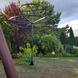 Metalowa konstrukcja z okręgami w ogrodzie, wsparcie dla roślin pnących, widok na trawnik i ozdobne krzewy, pochmurne niebo w tle.