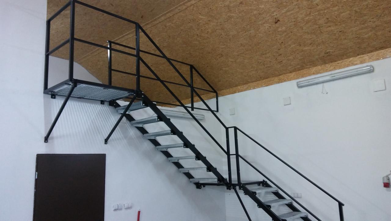 Metalowe schody z platformą, czarne barierki, surowy wystrój wnętrza, widok z dołu na konstrukcję stalową.