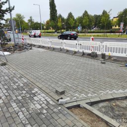 OL-KOP Usługi Koparką Mariusz Olczyk - Fragment chodnika z szarej kostki brukowej w trakcie układania, zabezpieczony białymi barierkami, widok na ulicę z przejeżdżającymi samochodami i drzewami w tle.