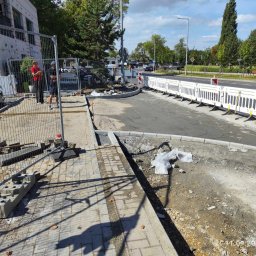 OL-KOP Usługi Koparką Mariusz Olczyk - Fragment remontowanej drogi z krawężnikami, zabezpieczony barierkami, z widocznymi pracownikami za ogrodzeniem i stertą kostki brukowej obok chodnika.