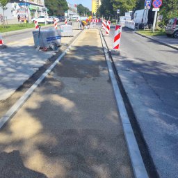 OL-KOP Usługi Koparką Mariusz Olczyk - Budowa chodnika przy ulicy: przygotowane podłoże z kruszywa, ułożone krawężniki, palety z kostką brukową i narzędzia w tle, robotnicy w oddali, znaki ostrzegawcze.