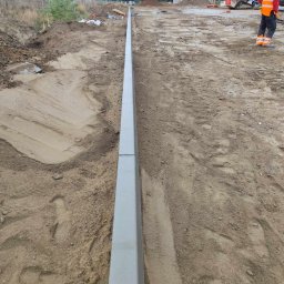 OL-KOP Usługi Koparką Mariusz Olczyk - Wyrównana linia betonowych obrzeży chodnikowych na placu budowy, widoczny pracownik w odblaskowej kamizelce i ciężki sprzęt w tle.