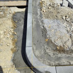 OL-KOP Usługi Koparką Mariusz Olczyk - Montaż obrzeży betonowych na podsypce cementowej, widoczne wykopane podłoże, żółta nić wyznaczająca linię, elementy kostki składowane obok.