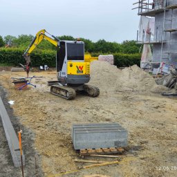 OL-KOP Usługi Koparką Mariusz Olczyk - Żółto-szara mini koparka Wacker Neuson na placu budowy, w tle budynek w trakcie budowy, obok leżące bloczki betonowe i ułożone obrzeża chodnika, pracownik w odblaskowej kamizelce.