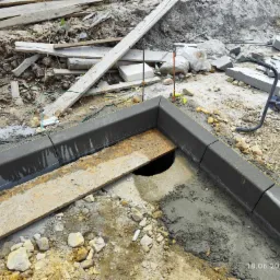 Montaż krawężnika betonowego, widoczna płyta OSB zakrywająca studzienkę, narzędzia budowlane, teren w trakcie przygotowania pod nawierzchnię, ziemia, piasek i gruz.