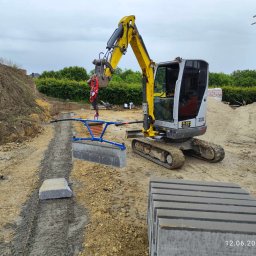 OL-KOP Usługi Koparką Mariusz Olczyk - Żółta mini koparka z niebieskim i szarym urządzeniem do podnoszenia kostki brukowej, przygotowany wykop pod kostkę, stos kostki brukowej, piasek i budynek w tle