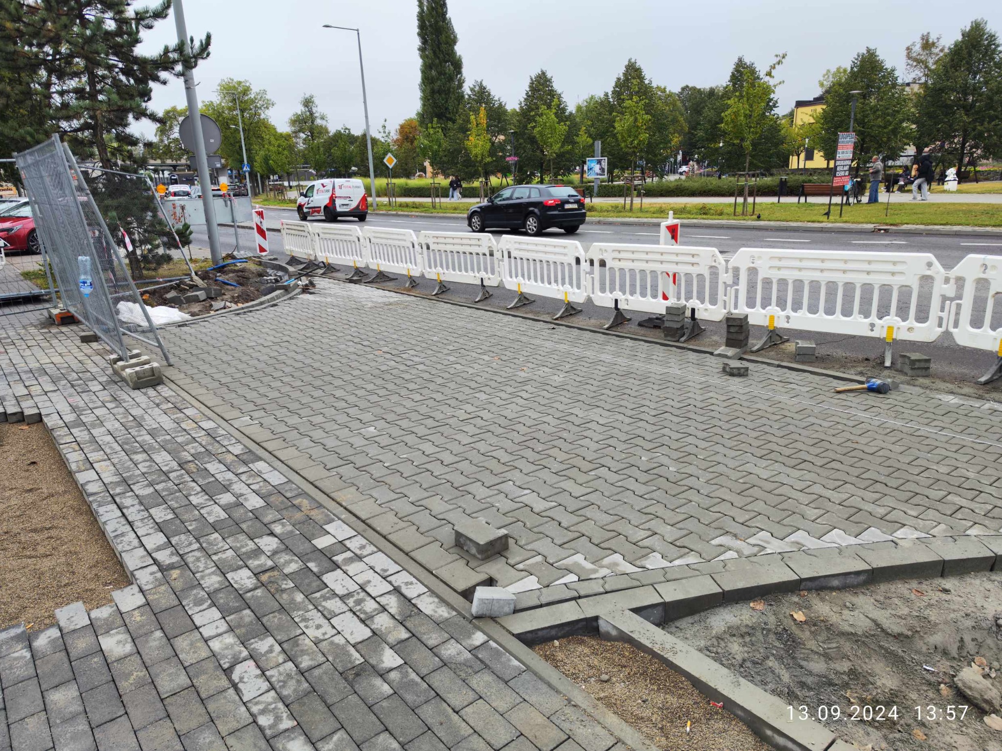 Fragment chodnika z szarej kostki brukowej w trakcie układania, zabezpieczony białymi barierkami, widok na ulicę z przejeżdżającymi samochodami i drzewami w tle.