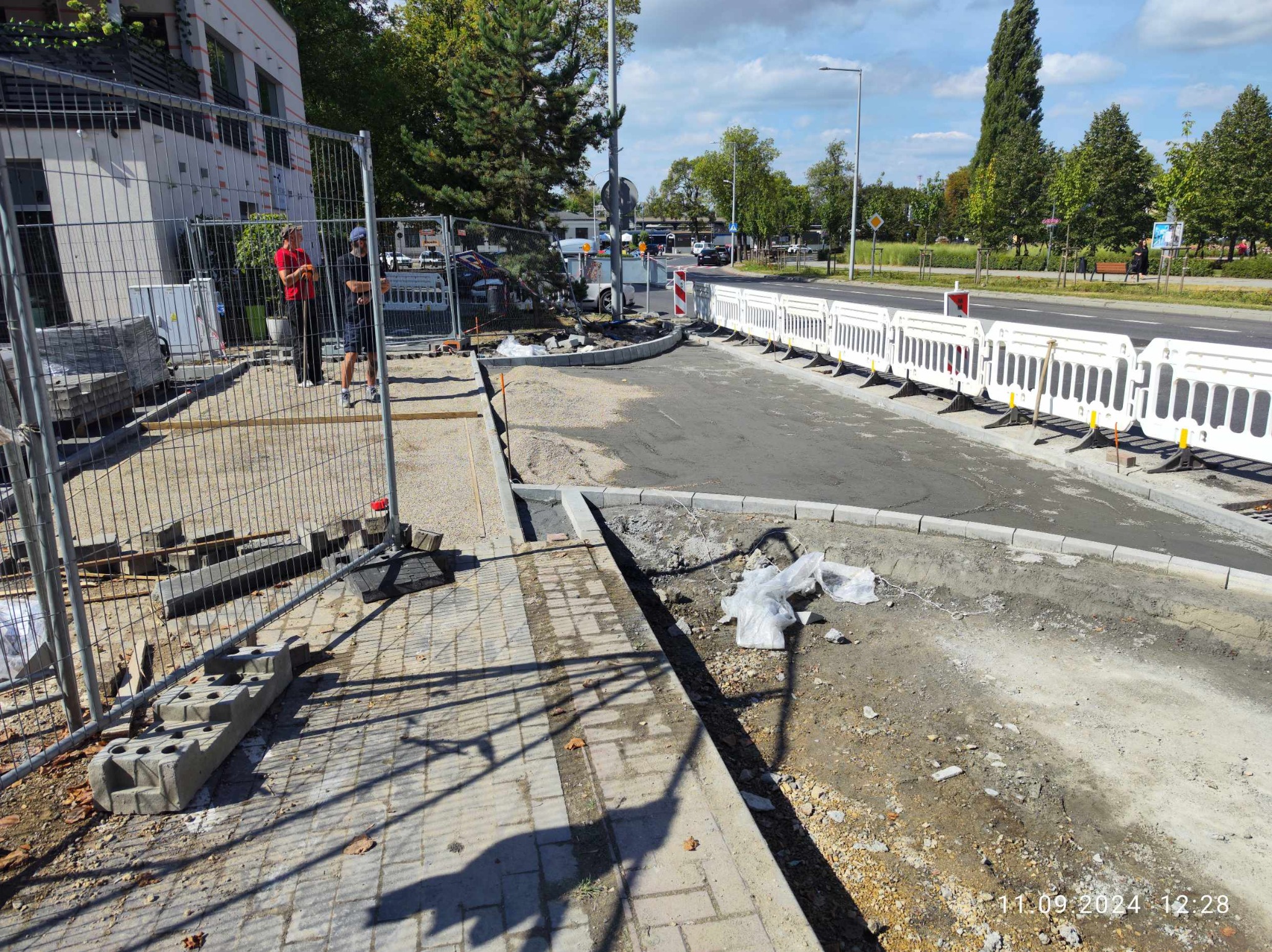Fragment remontowanej drogi z krawężnikami, zabezpieczony barierkami, z widocznymi pracownikami za ogrodzeniem i stertą kostki brukowej obok chodnika.