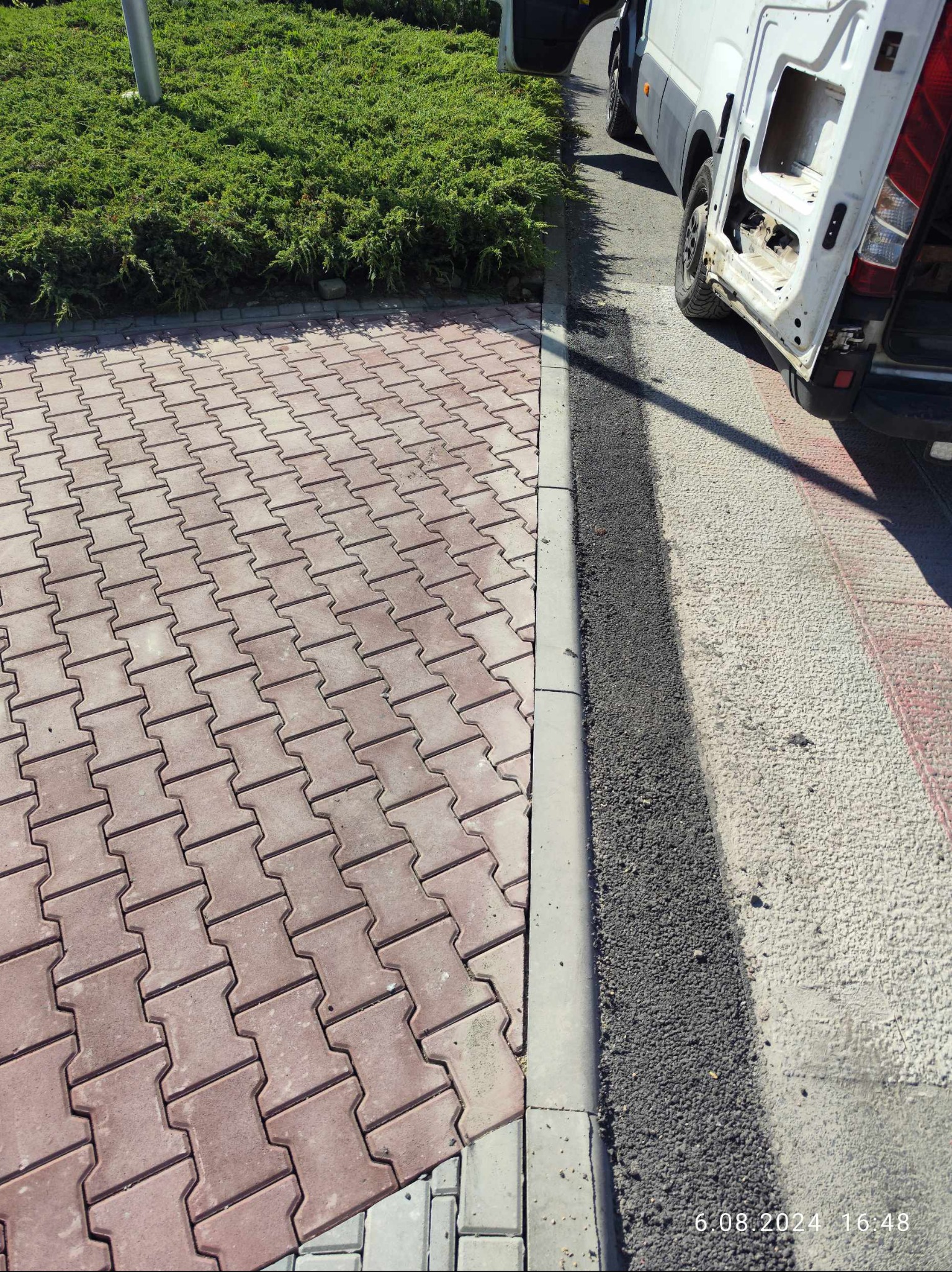 Częściowo ułożona, czerwona kostka brukowa w kształcie litery H, oddzielona krawężnikiem od świeżo wylanego asfaltu, obok zaparkowany biały samochód dostawczy z otwartymi drzwiami.