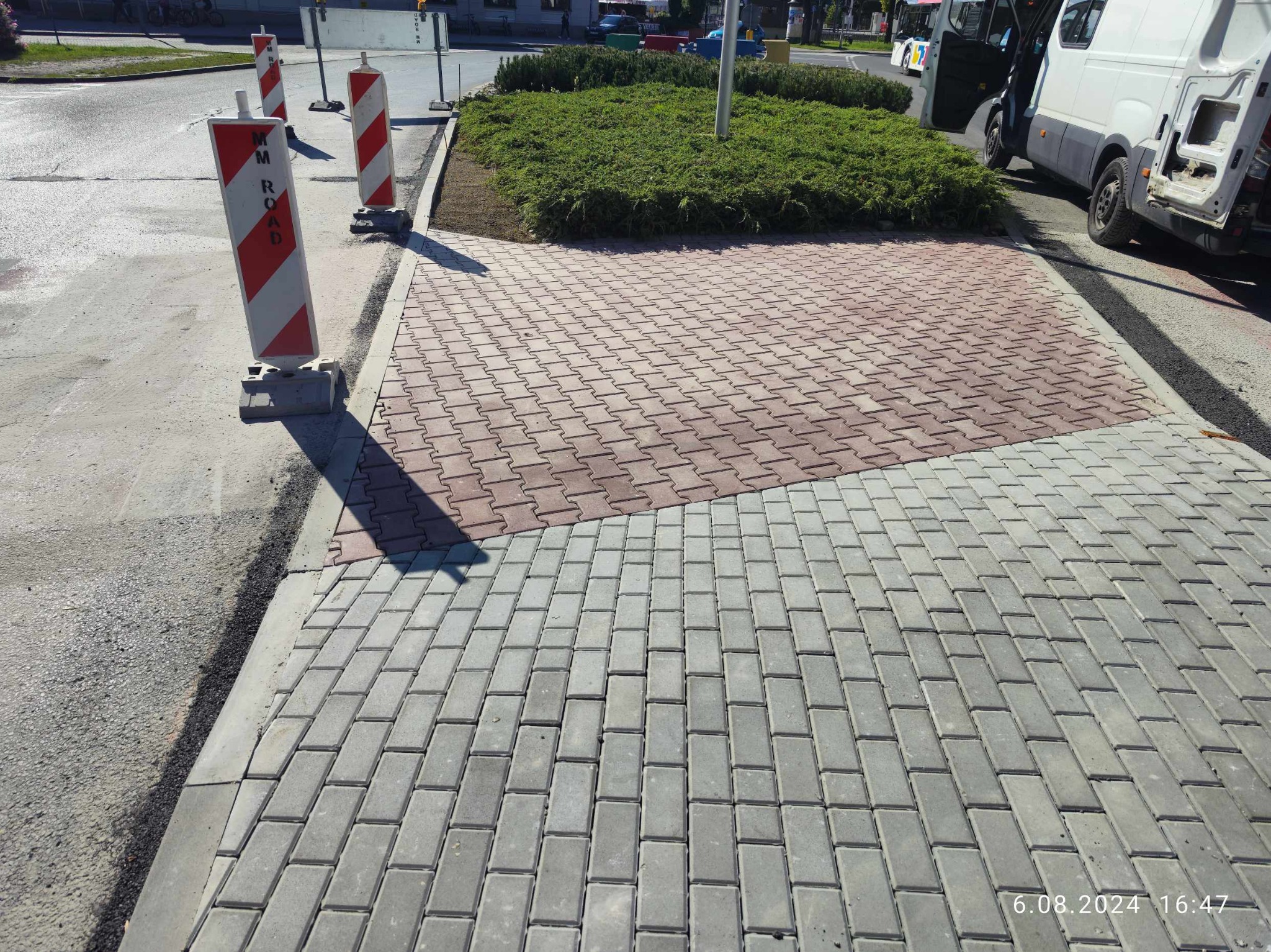 Fragment nowej nawierzchni z kostki brukowej w dwóch kolorach, oddzielającej jezdnię od pasa zieleni, z widocznymi znakami drogowymi i samochodem dostawczym w tle.