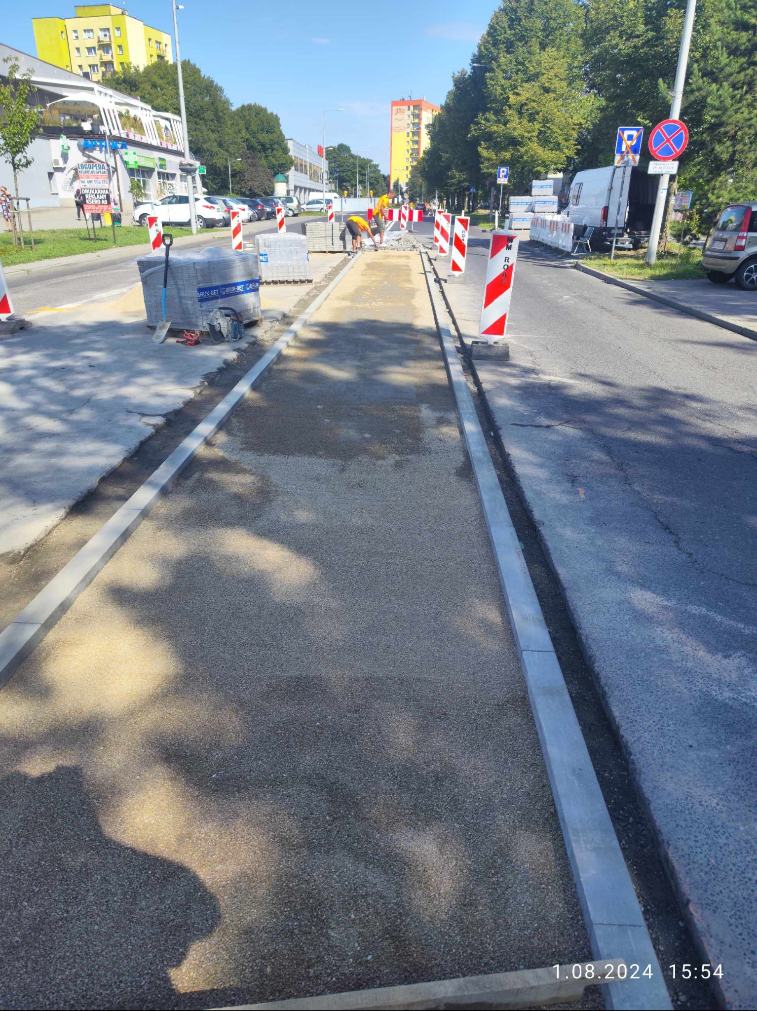 Budowa chodnika przy ulicy: przygotowane podłoże z kruszywa, ułożone krawężniki, palety z kostką brukową i narzędzia w tle, robotnicy w oddali, znaki ostrzegawcze.