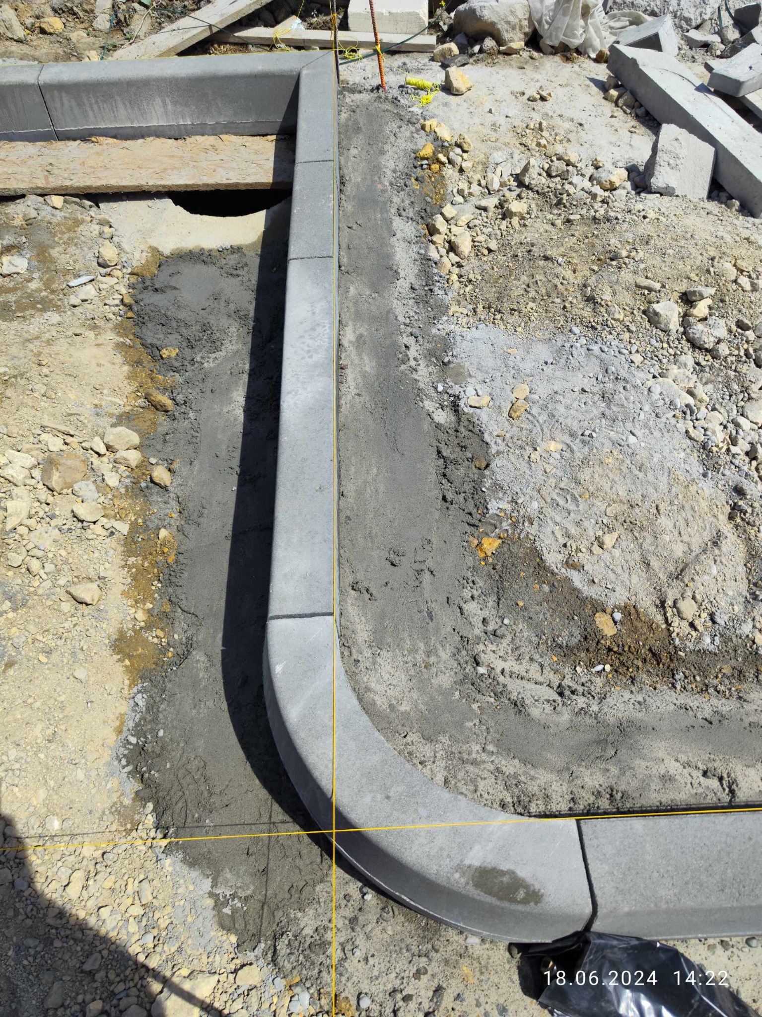 Montaż obrzeży betonowych na podsypce cementowej, widoczne wykopane podłoże, żółta nić wyznaczająca linię, elementy kostki składowane obok.