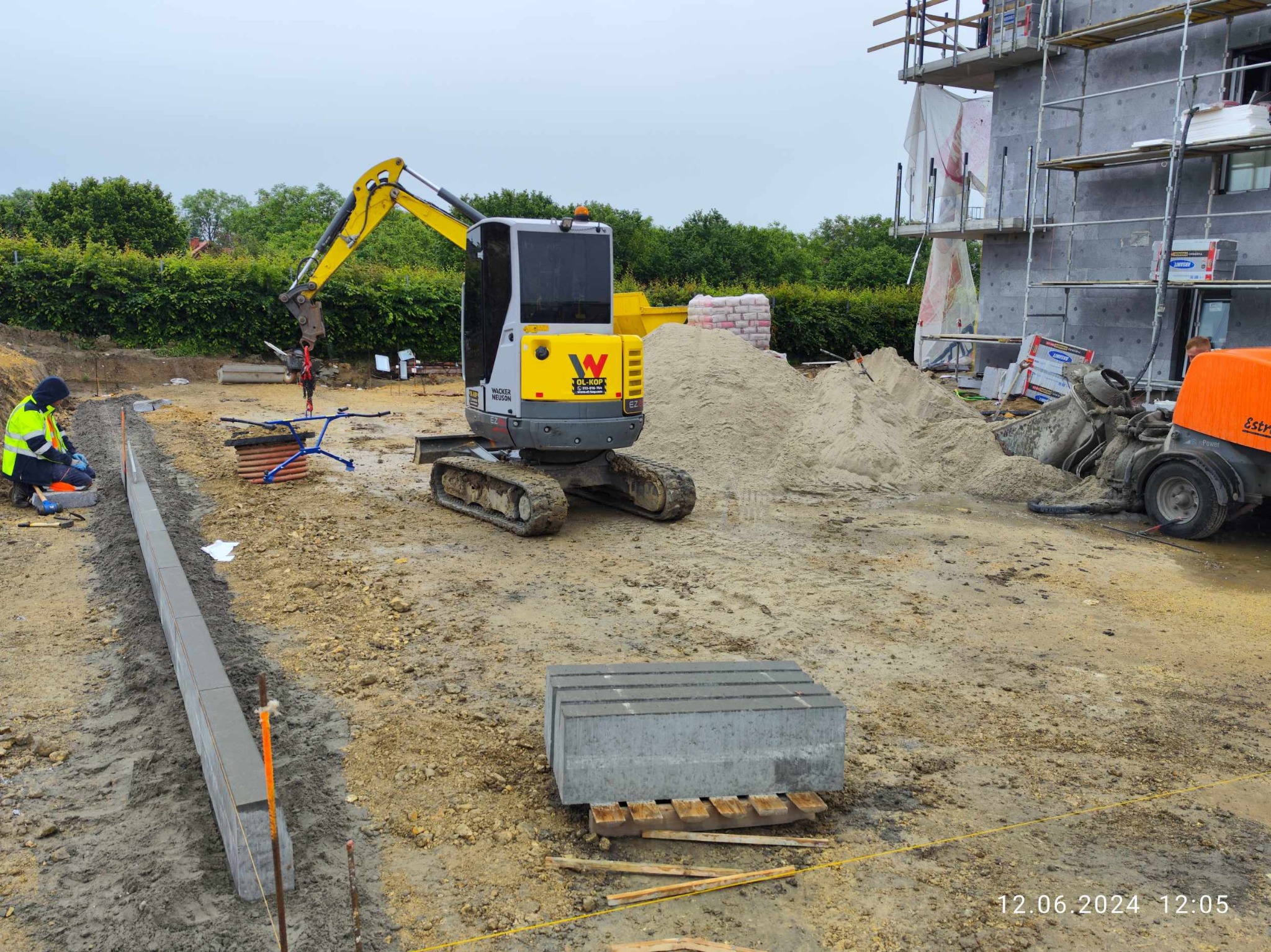 Żółto-szara mini koparka Wacker Neuson na placu budowy, w tle budynek w trakcie budowy, obok leżące bloczki betonowe i ułożone obrzeża chodnika, pracownik w odblaskowej kamizelce.
