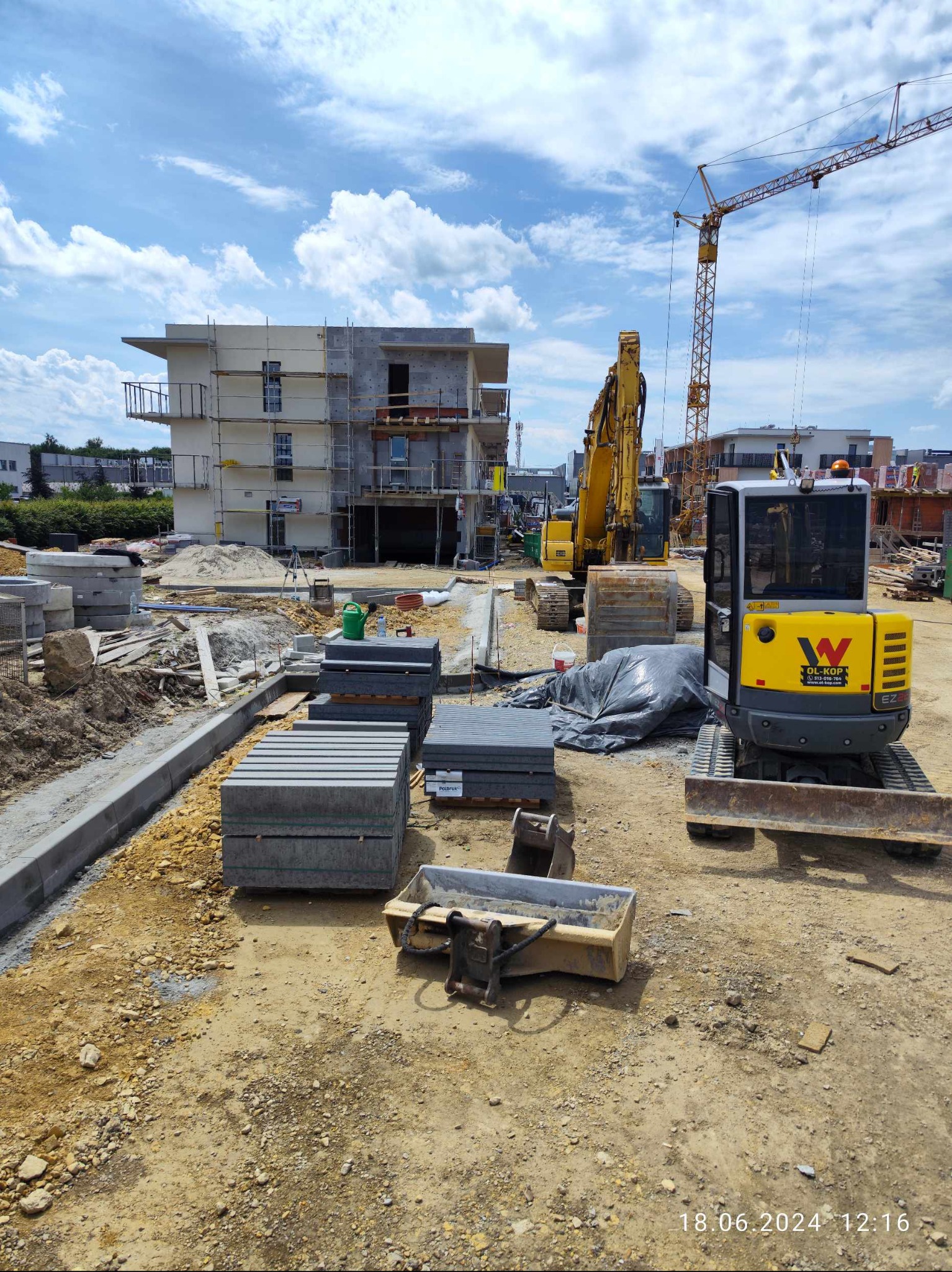 Plac budowy z wykopem, paletami kostki brukowej, mini koparką z logo firmy i budynkiem w trakcie budowy w tle, widoczny żuraw budowlany.
