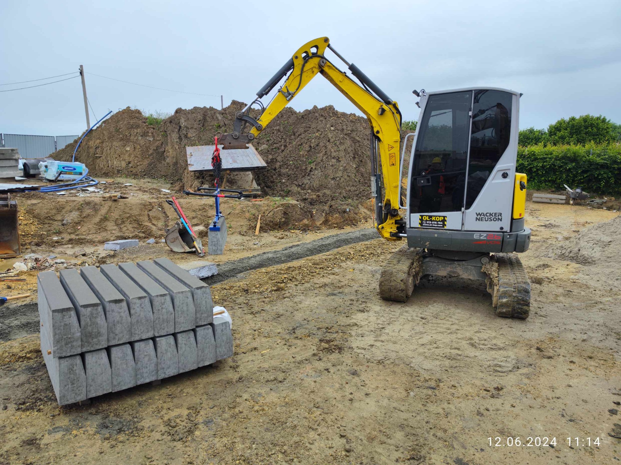 Żółto-szara mini koparka Wacker Neuson z operatorem w kabinie, przygotowuje teren pod ułożenie obrzeży chodnikowych, widoczny wykop i stos obrzeży.