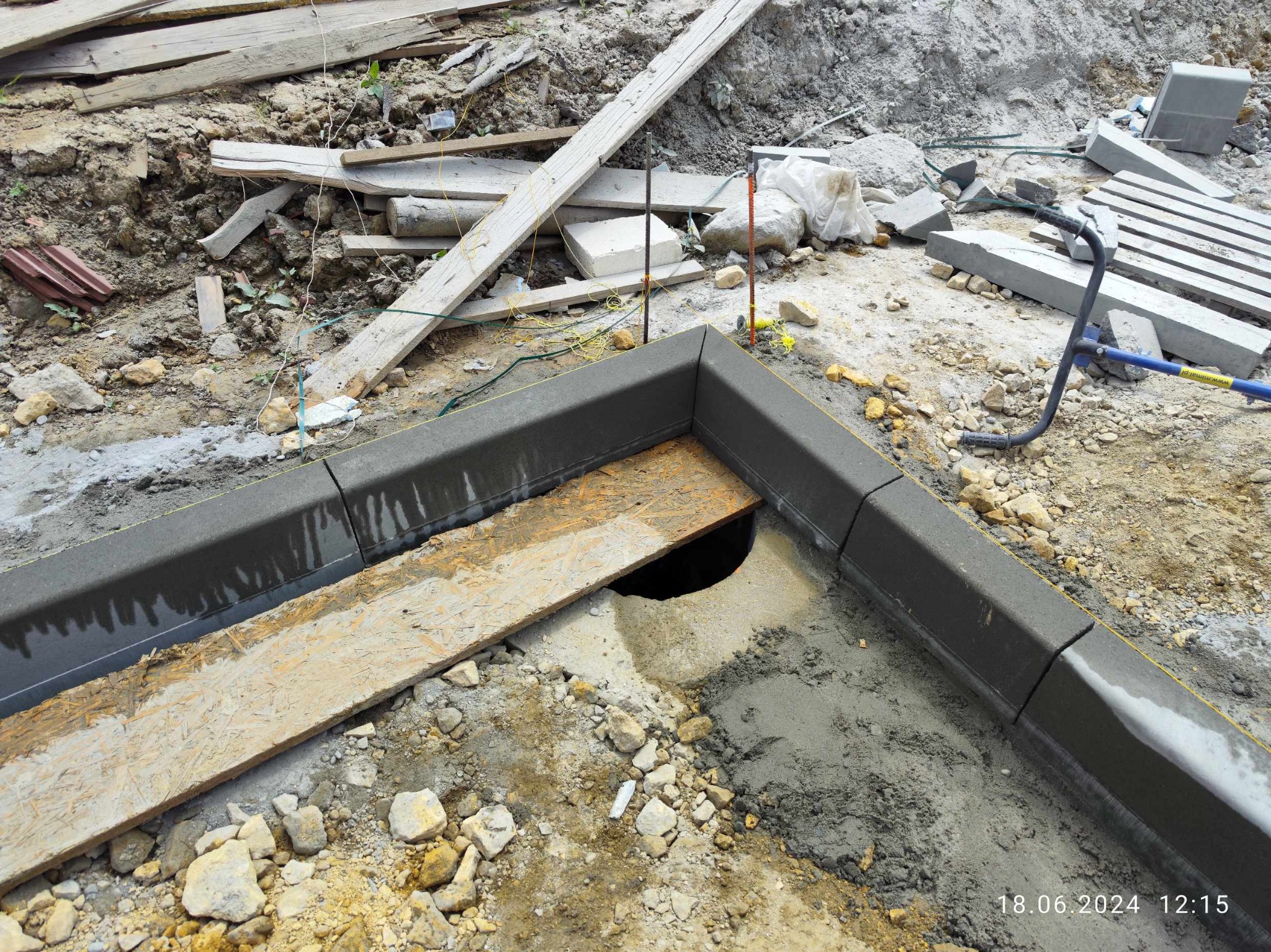 Montaż krawężnika betonowego, widoczna płyta OSB zakrywająca studzienkę, narzędzia budowlane, teren w trakcie przygotowania pod nawierzchnię, ziemia, piasek i gruz.