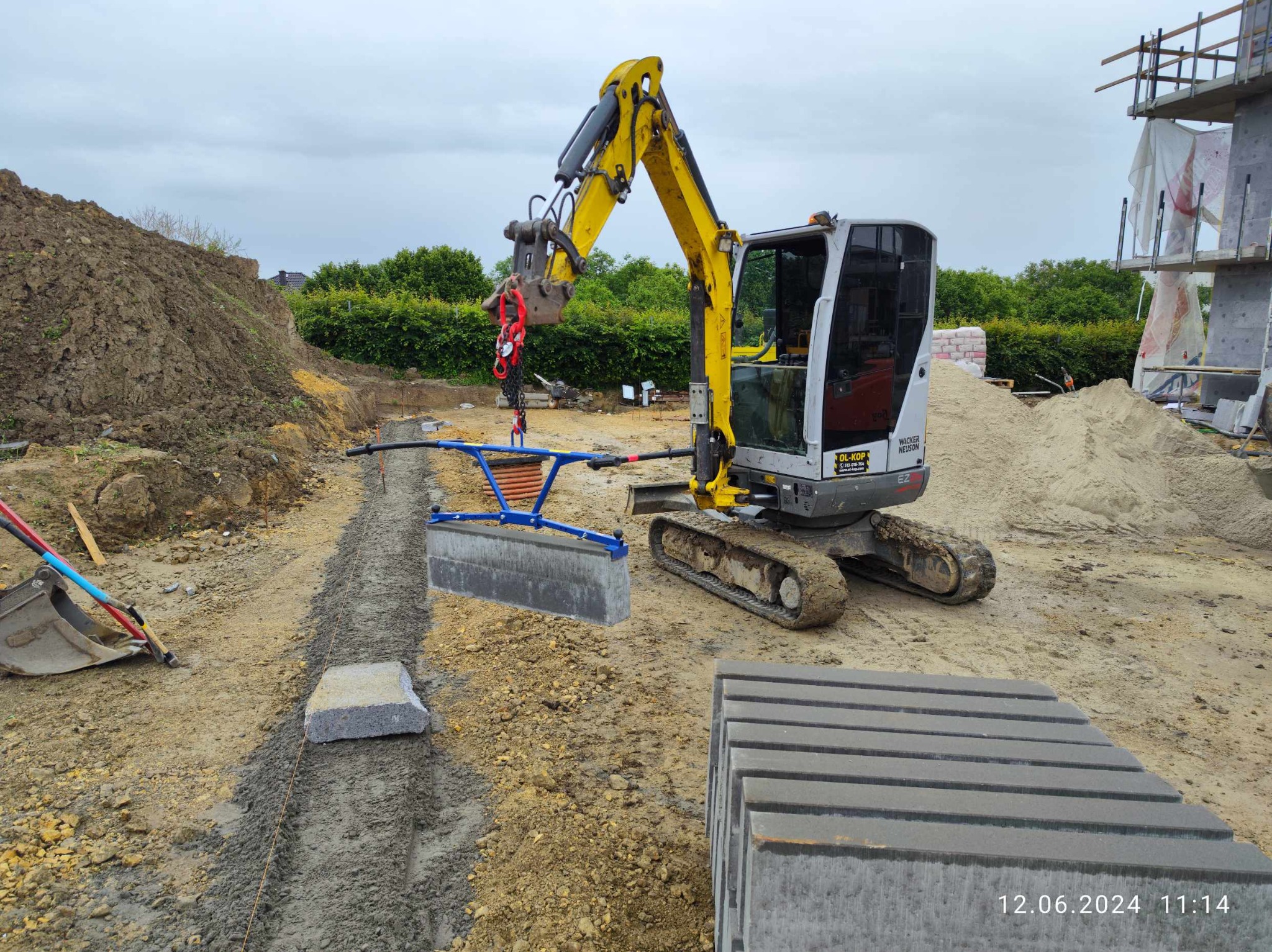 Żółta mini koparka z niebieskim i szarym urządzeniem do podnoszenia kostki brukowej, przygotowany wykop pod kostkę, stos kostki brukowej, piasek i budynek w tle