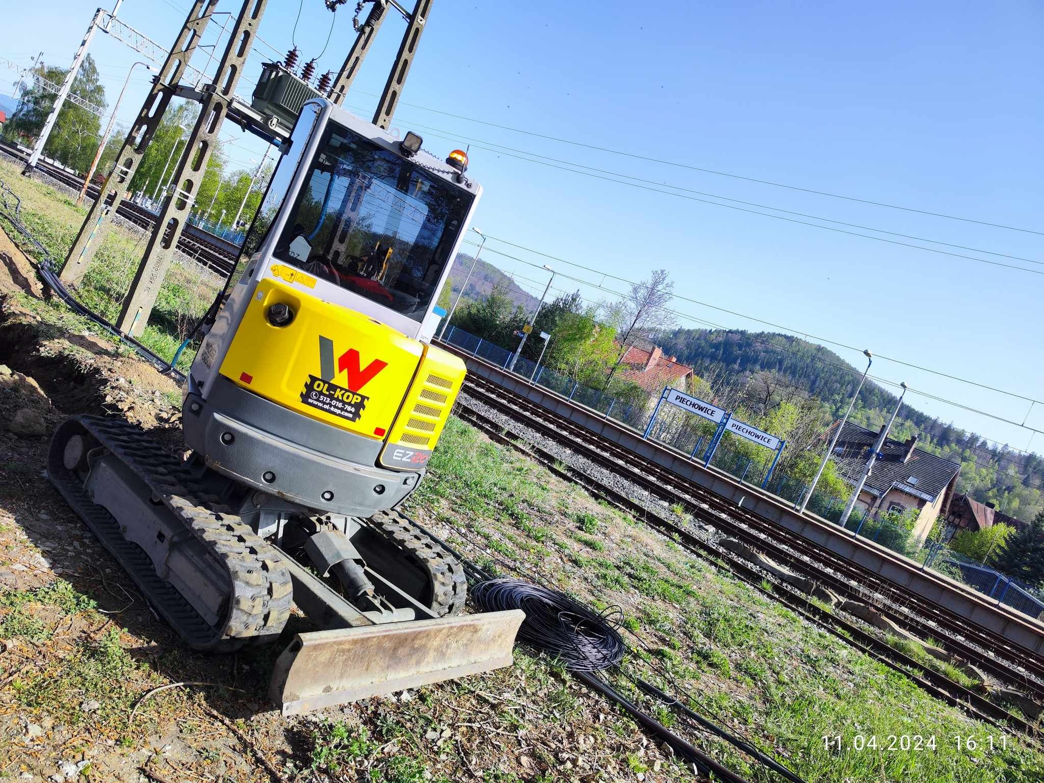 Żółto-szara mini koparka Wacker Neuson przy wykopie w pobliżu torów kolejowych i słupów energetycznych, Piechowice w tle.