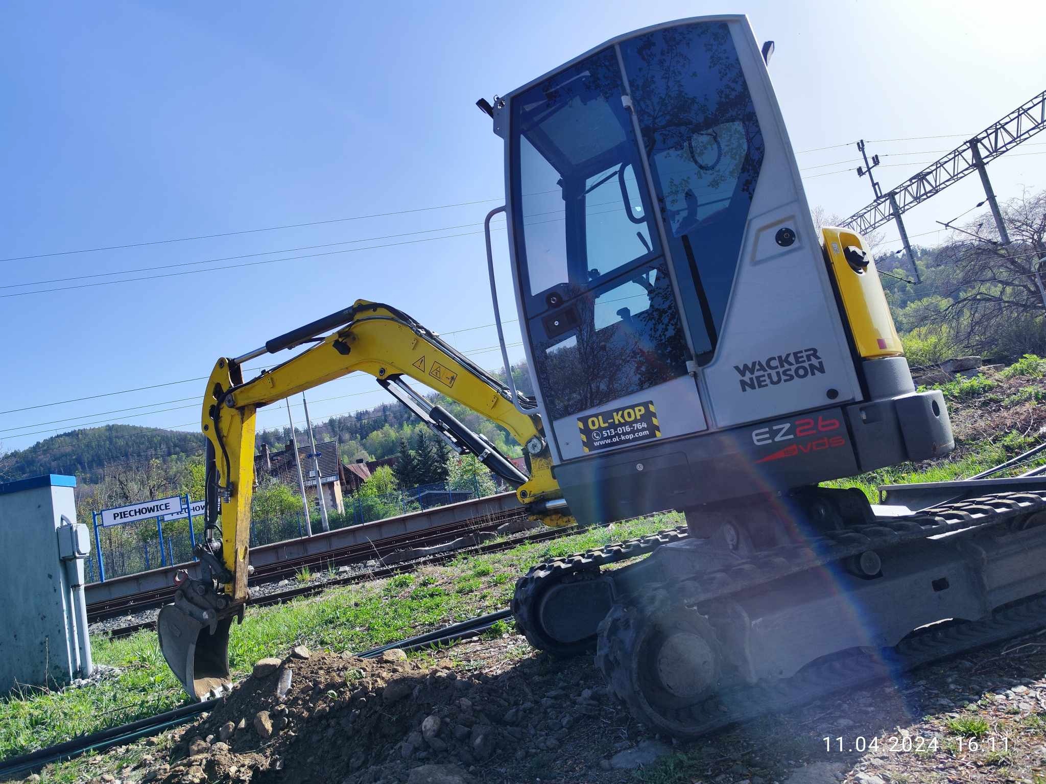 Żółto-szara minikoparka Wacker Neuson EZ26 przy torach kolejowych w słoneczny dzień, widoczny znak stacji Piechowice i częściowo odsłonięty kabel w ziemi.