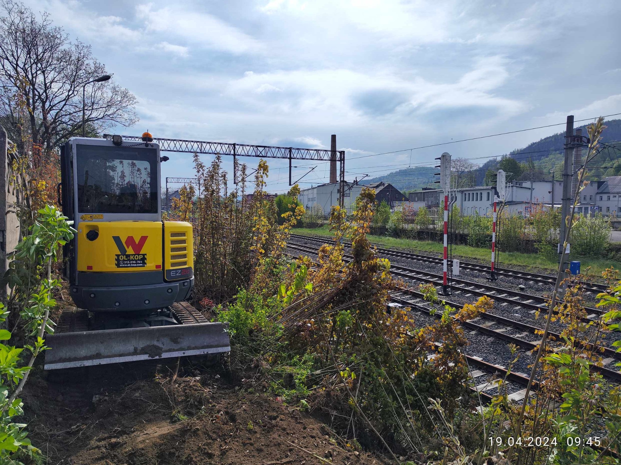 Żółta mini koparka OL-KOP EZ26 na tle torów kolejowych i zabudowań przemysłowych, przygotowująca teren pod instalacje, widoczne świeżo naruszone podłoże.