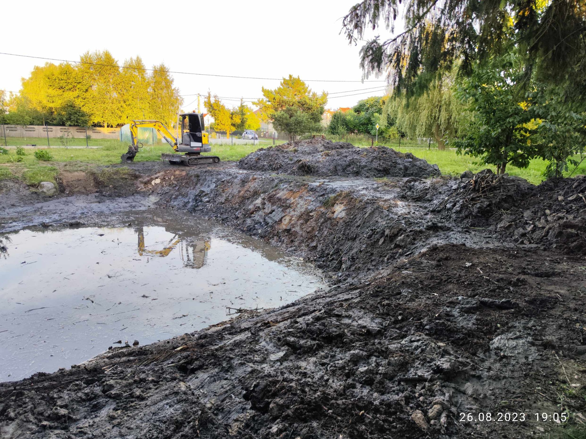 Żółto-szara minikoparka odbija się w wodzie wykopanego stawu, w tle drzewa w popołudniowym słońcu, hałdy ziemi na pierwszym planie.