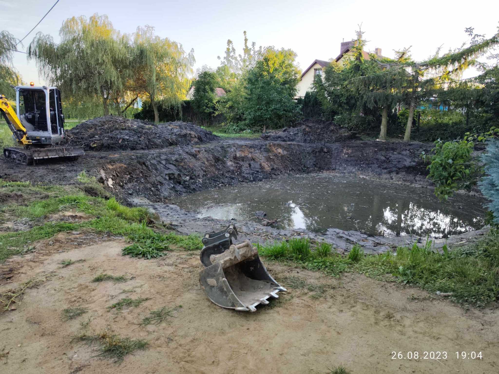 Wyrobisko wypełnione wodą na działce, z minikoparką w tle i łyżką koparki na pierwszym planie, widoczne ślady pracy ziemnej.