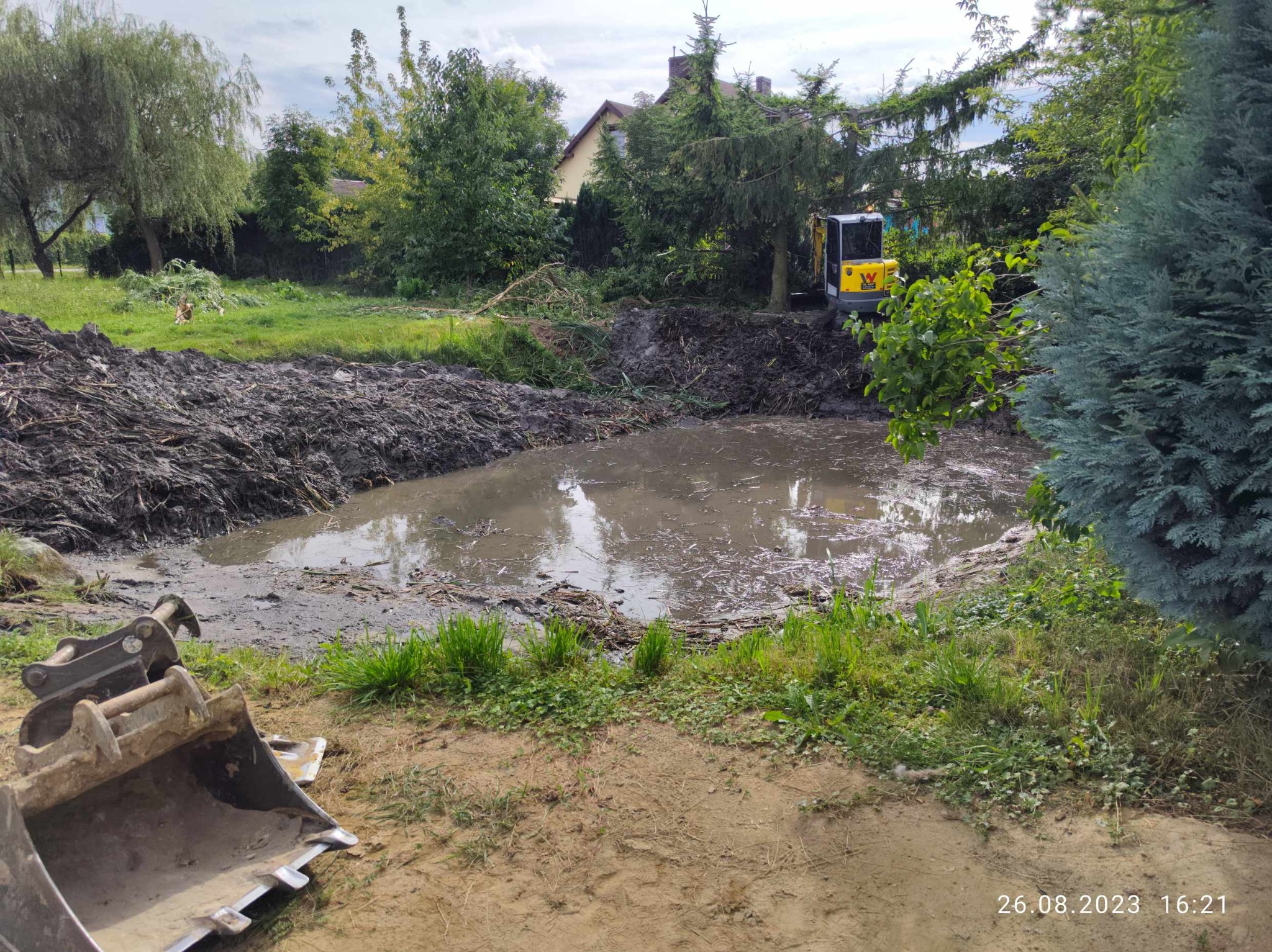 Wyrobisko wypełnione wodą i błotem, na brzegu sterta ziemi i trawy, w tle żółta minikoparka i zieleń drzew, widoczny fragment łyżki koparki na pierwszym planie.