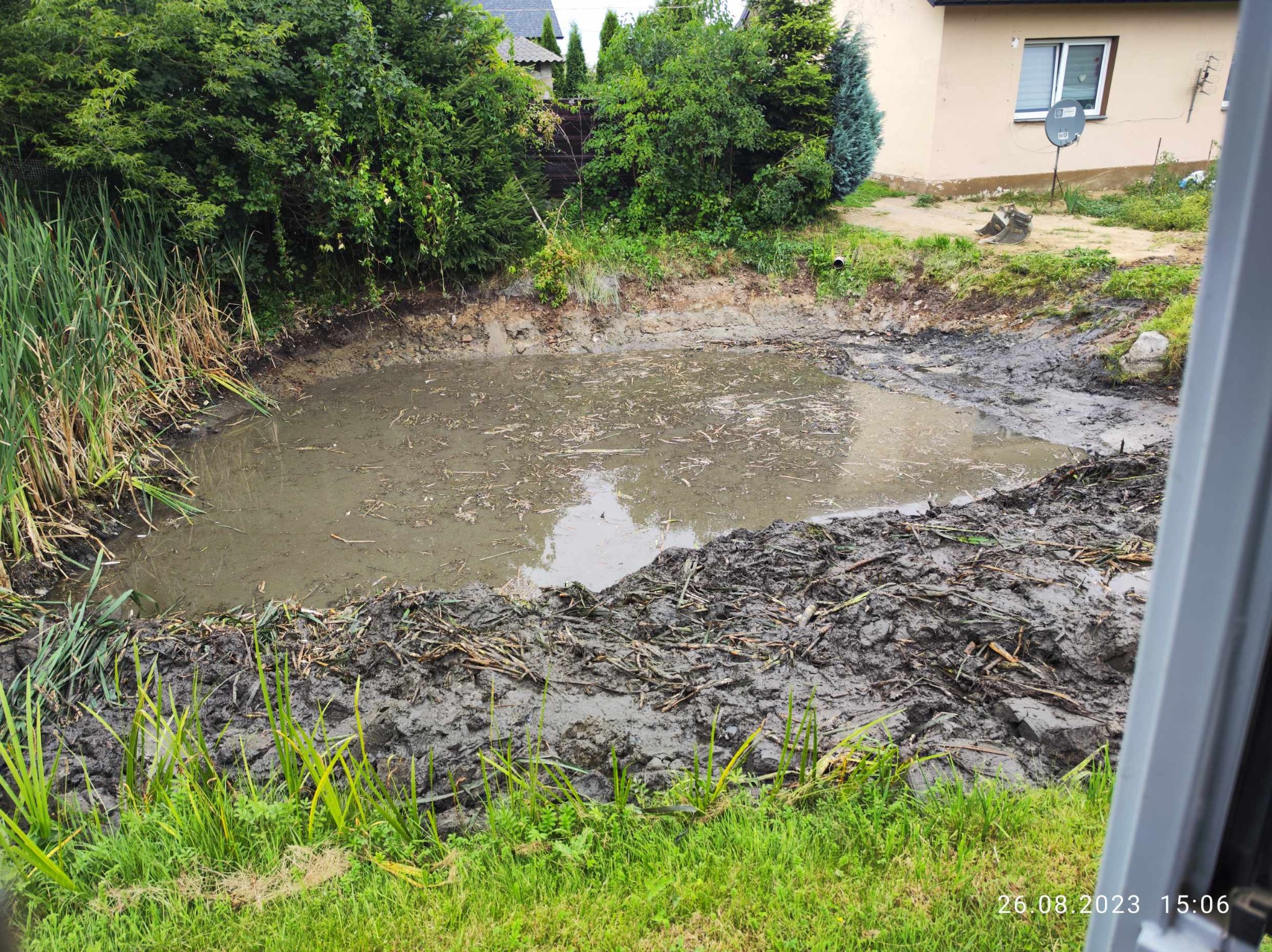 Wyrobisko wypełnione wodą i błotem, otoczone bujną zielenią, w tle budynek z anteną satelitarną.