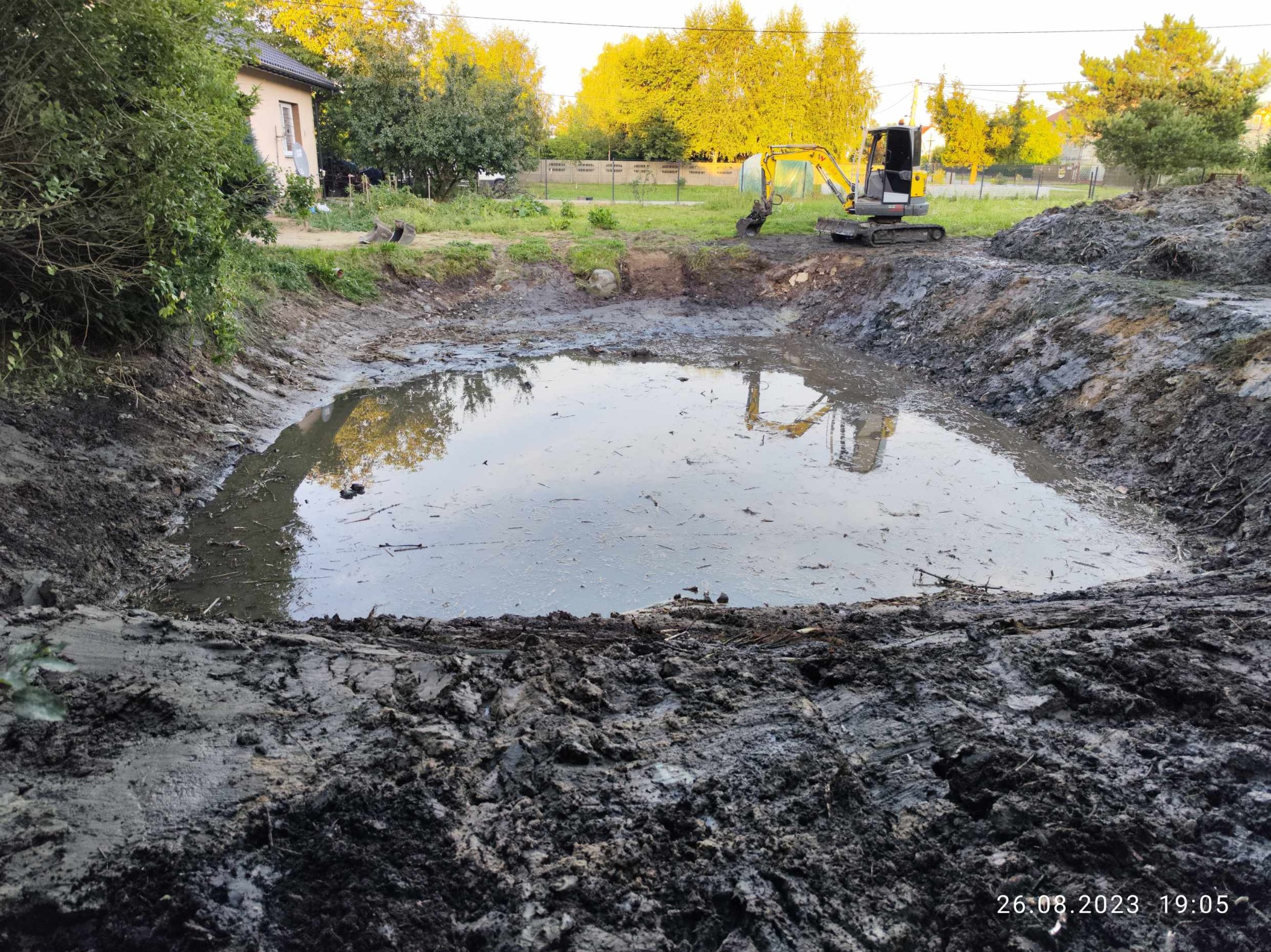 Wykonany wykop pod oczko wodne, częściowo wypełniony wodą, z widoczną małą koparką na brzegu i domem w tle, wykonany w pochmurny dzień.