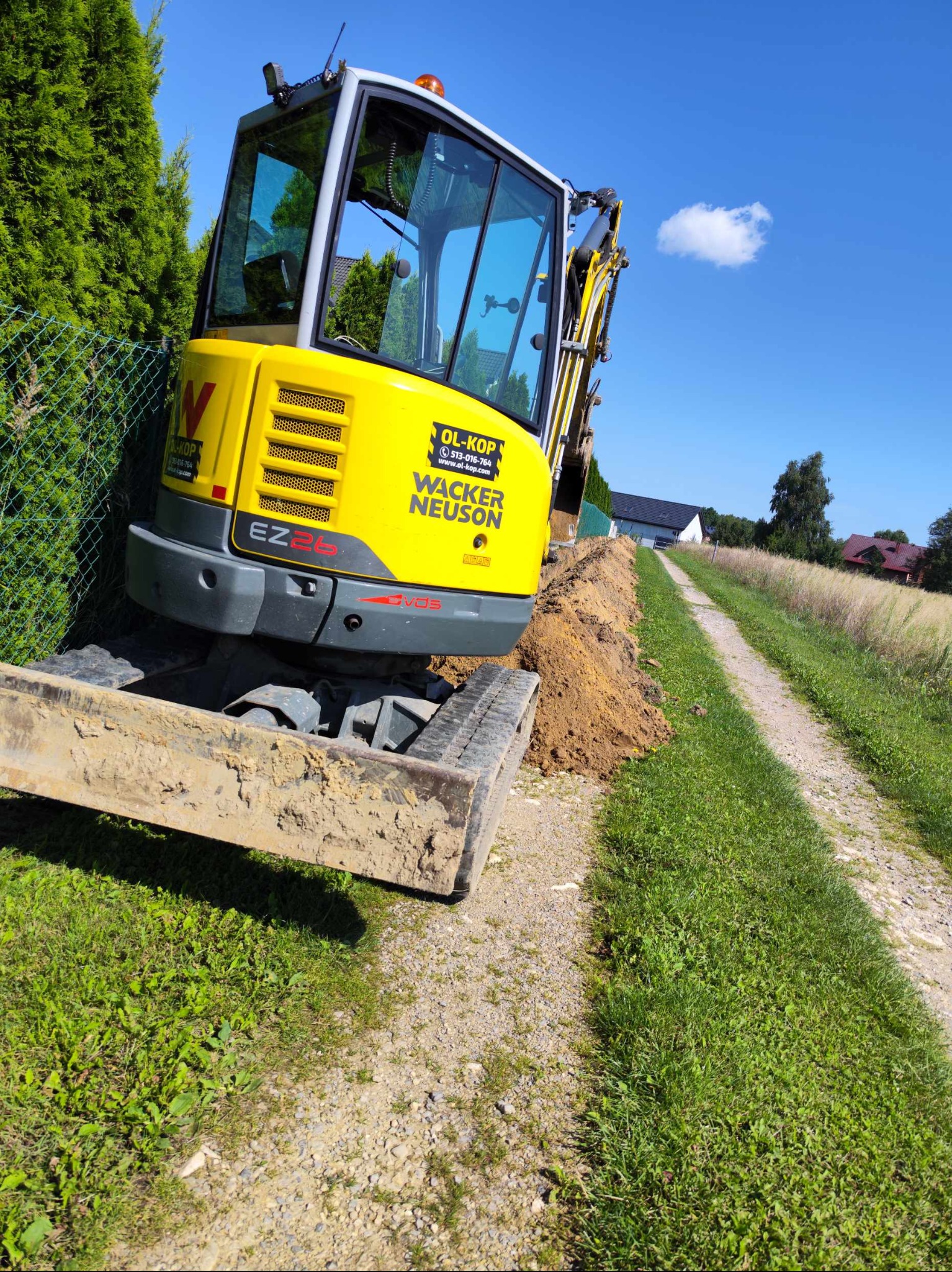 Żółta koparka Wacker Neuson EZ26 na grząskiej drodze, obok wykopu ziemnego i zielonego żywopłotu, błękitne niebo z pojedynczą chmurą w tle.