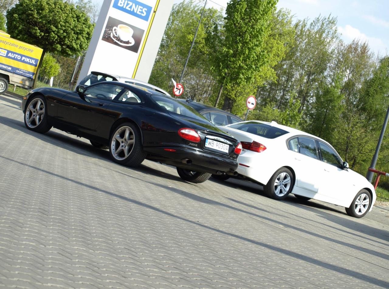 Czarny Jaguar XK zaparkowany obok białego BMW na parkingu z kostki brukowej, widoczny znak 'Biznes' w tle.