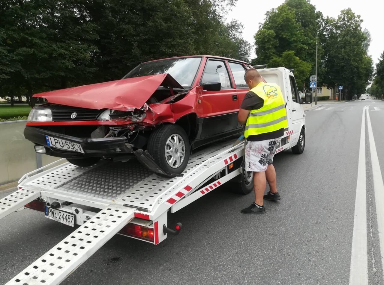 Czerwony uszkodzony samochód marki FSO Polonez na białej lawecie, zabezpieczany przez pracownika w kamizelce odblaskowej z logo firmy 'freecar'.