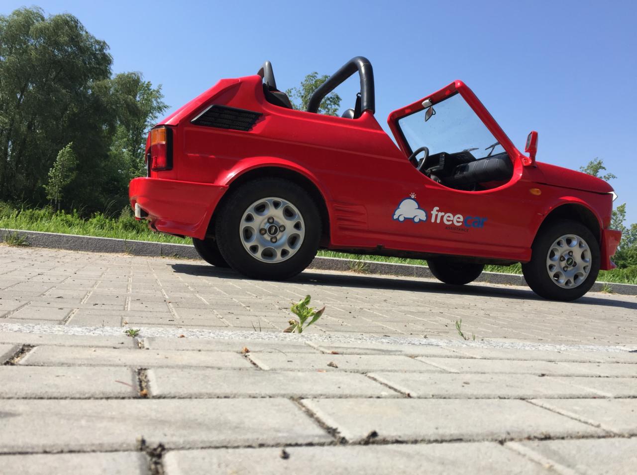 Czerwony, nietypowy kabriolet z logotypem 'free car assistance' zaparkowany na brukowanym parkingu, z młodą rośliną wyrastającą spomiędzy kostek.
