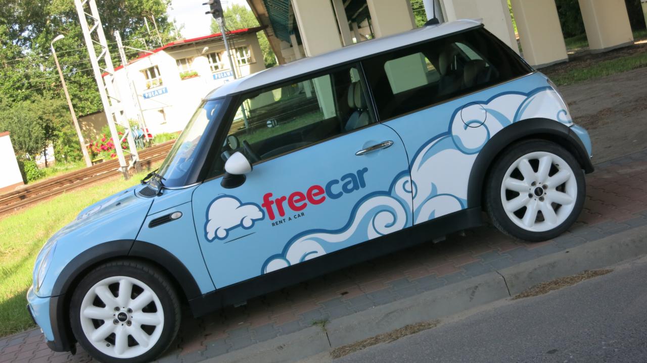 Jasnoniebieski Mini Cooper z białymi felgami i grafiką chmur oraz logo 'freecar rent a car' zaparkowany na chodniku, w tle budynek stacji kolejowej i tory.