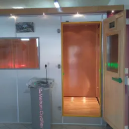 Komora krioterapeutyczna z otwartymi drzwiami, widoczna drewniana podłoga wewnątrz, obok stanowisko kontrolne z mikrofonem i nazwą 'Metrum CryoFlex'.