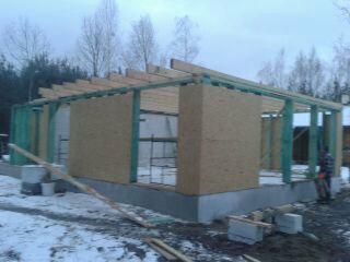 Wznoszenie konstrukcji budynku z płyt OSB na fundamencie betonowym, widoczne elementy szkieletu drewnianego, zimowy krajobraz w tle.