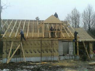 Wznoszenie konstrukcji dachu budynku w technologii szkieletowej, widoczni pracownicy na rusztowaniach, pochmurne niebo w tle.